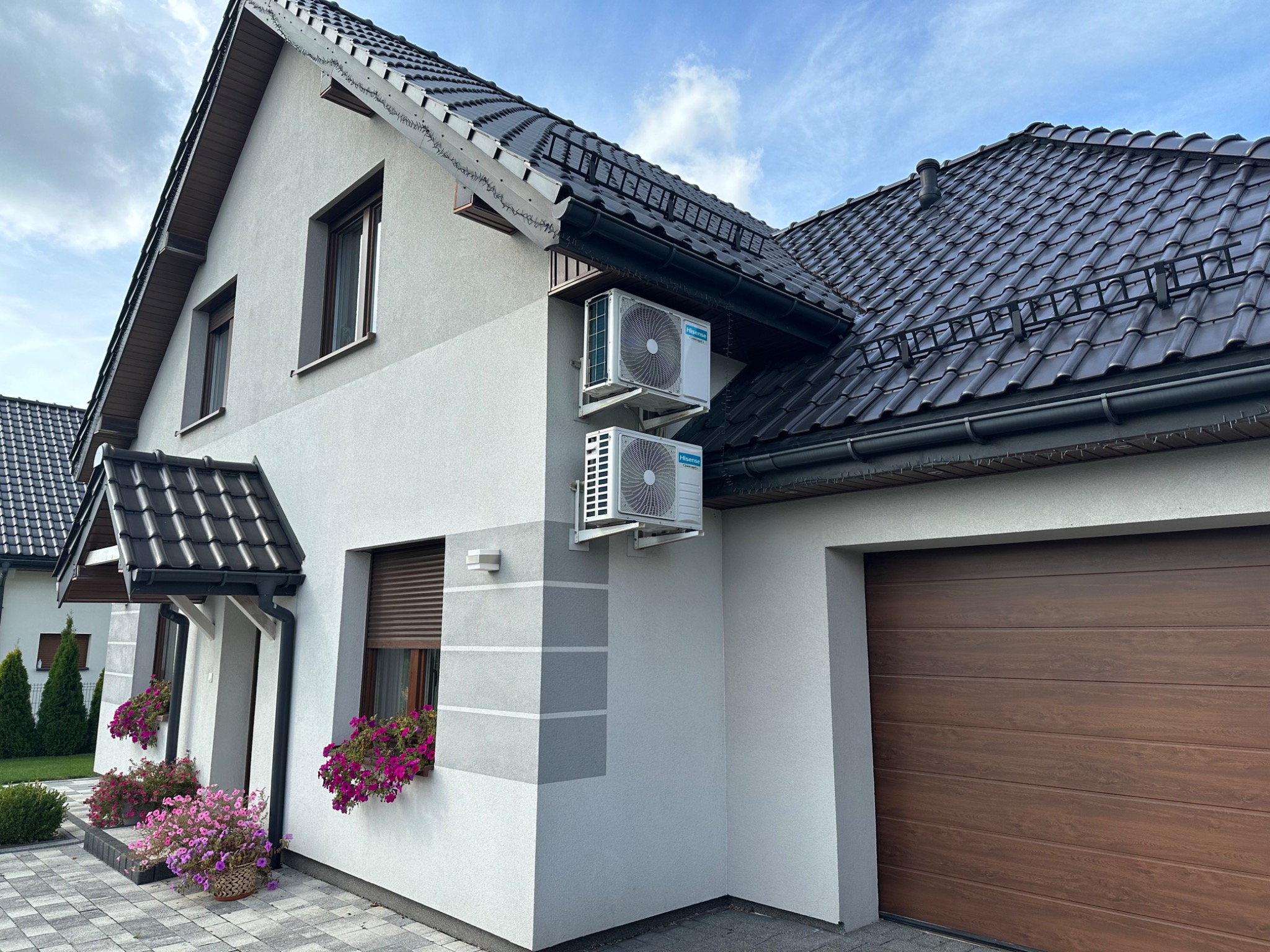 Dwa białe klimatyzatory zamontowane na elewacji domu jednorodzinnego z brązowym dachem i garażem, widoczne kwiaty w oknach.