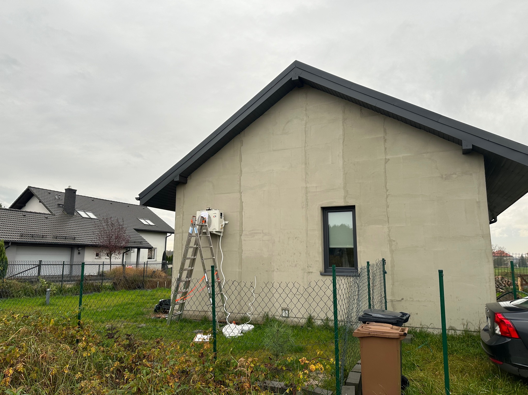 Montaż zewnętrznej jednostki klimatyzacji na ścianie domu jednorodzinnego, widoczna drabina i przewody, w tle drugi dom i pochmurne niebo.