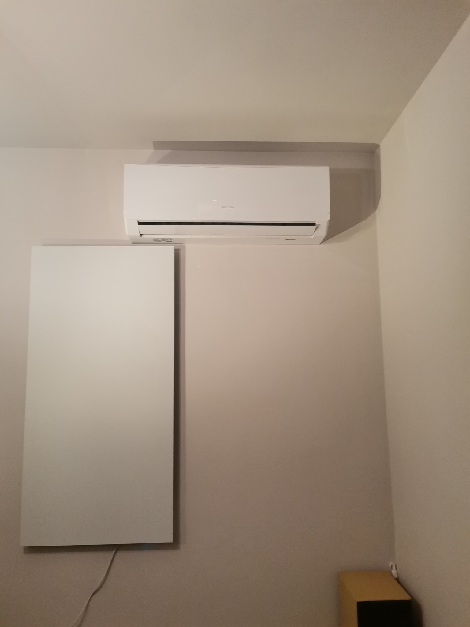 Biały klimatyzator ścienny zamontowany w rogu pokoju, obok panelu grzewczego z widocznym kablem zasilającym.