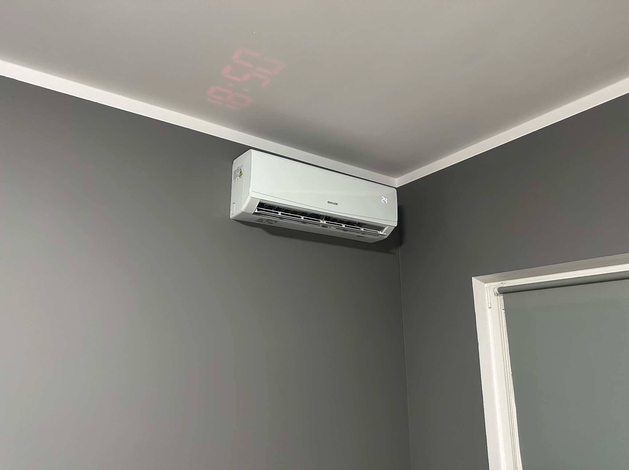 Biała klimatyzacja Sinclair zamontowana na szarej ścianie w pomieszczeniu, z wyświetlonym na suficie czerwonym zegarem cyfrowym.