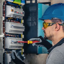 pomiary.mobi - Elektryk w niebieskim kasku i okularach ochronnych, z czerwoną śrubokrętem, pracuje przy rozdzielnicy elektrycznej z bezpiecznikami i przewodami, w rękawicach roboczych.