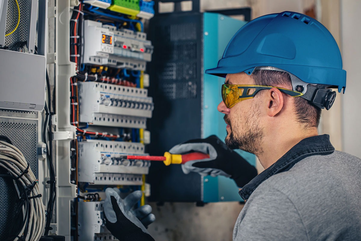 Elektryk w niebieskim kasku i okularach ochronnych, z czerwoną śrubokrętem, pracuje przy rozdzielnicy elektrycznej z bezpiecznikami i przewodami, w rękawicach roboczych.