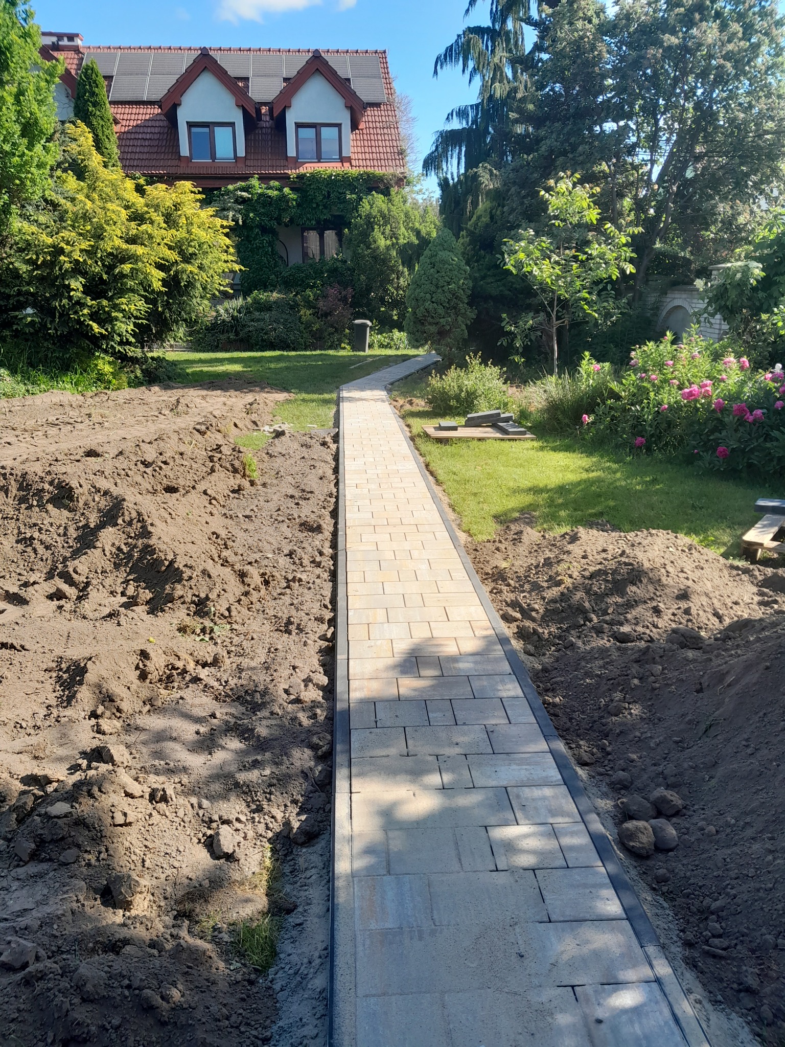Wąska ścieżka z jasnoszarej kostki brukowej prowadząca przez ogród w kierunku domu z czerwonym dachem i panelami słonecznymi, widoczne przygotowane podłoże i trawnik z kwiatami.