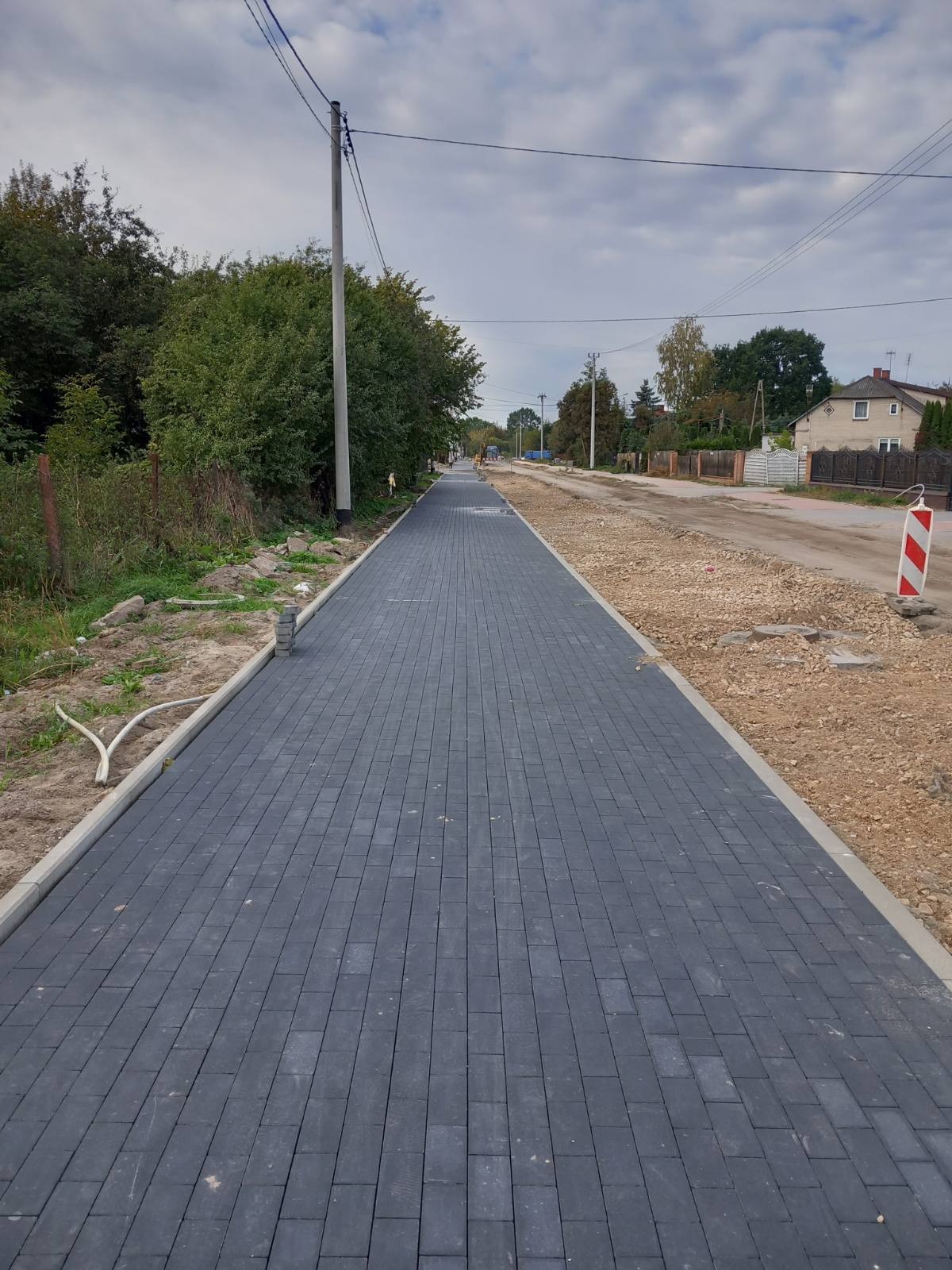 Długa, nowo ułożona, ciemnoszara kostka brukowa na chodniku, widok w perspektywie, z widocznym przygotowaniem terenu pod dalsze prace po prawej stronie i słupem elektrycznym w tle.