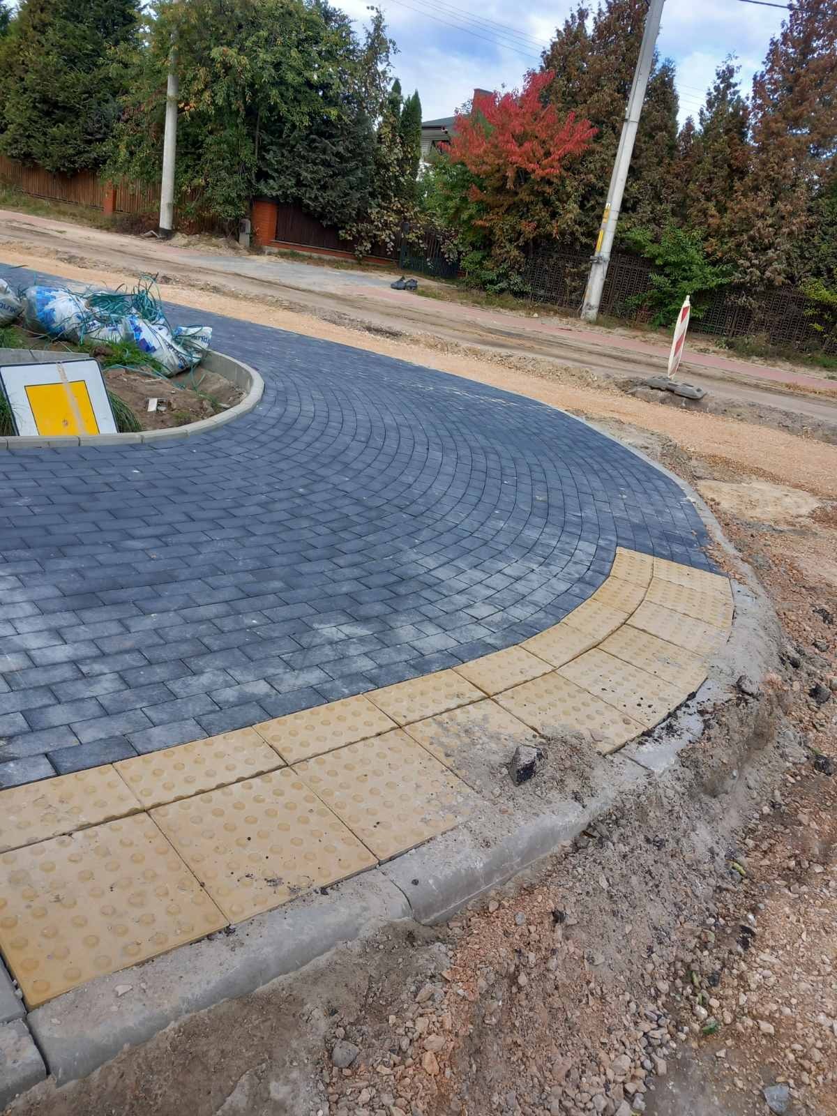 Niebieska kostka brukowa ułożona w łuk na skrzyżowaniu drogi, z żółtymi płytami dla niewidomych przy krawężniku, w tle worki z materiałem i znak drogowy.