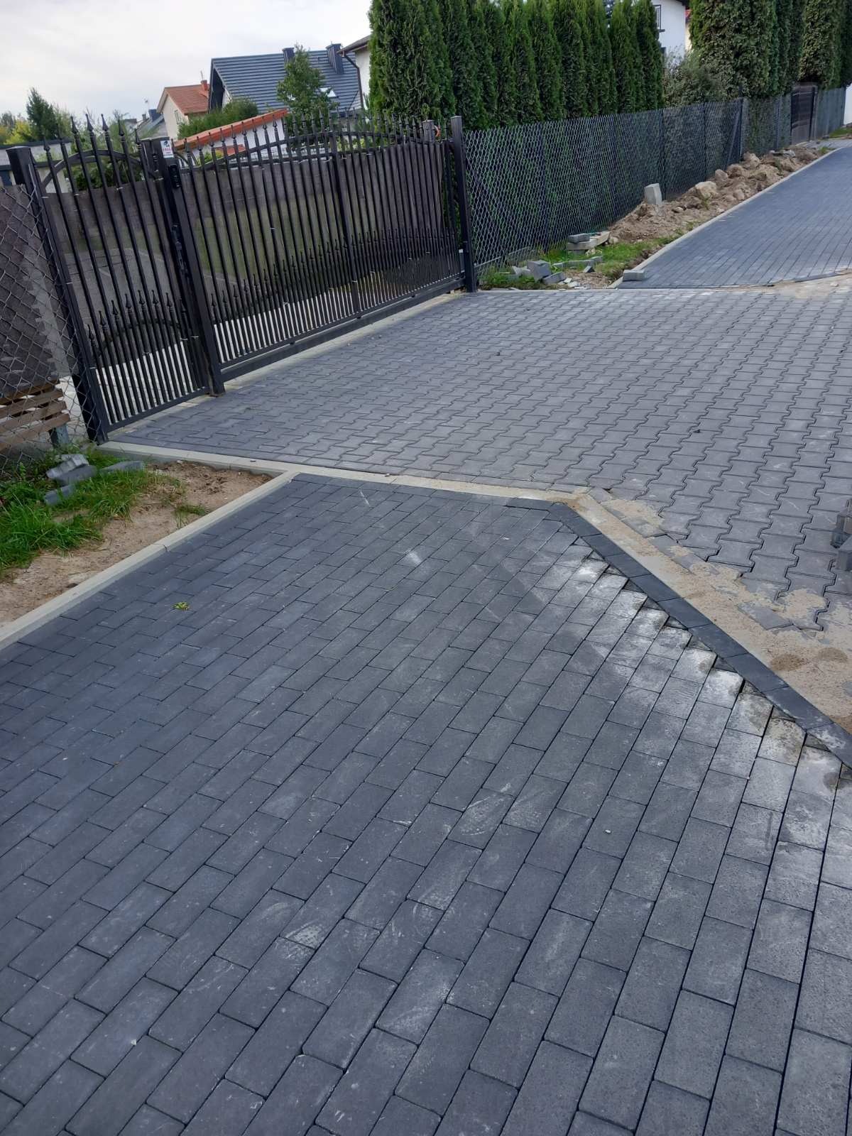 Ułożona, szara kostka brukowa przed posesją z czarną, metalową bramą i ogrodzeniem z siatki. Widoczne są również elementy zieleni i fragmenty budynków w tle.
