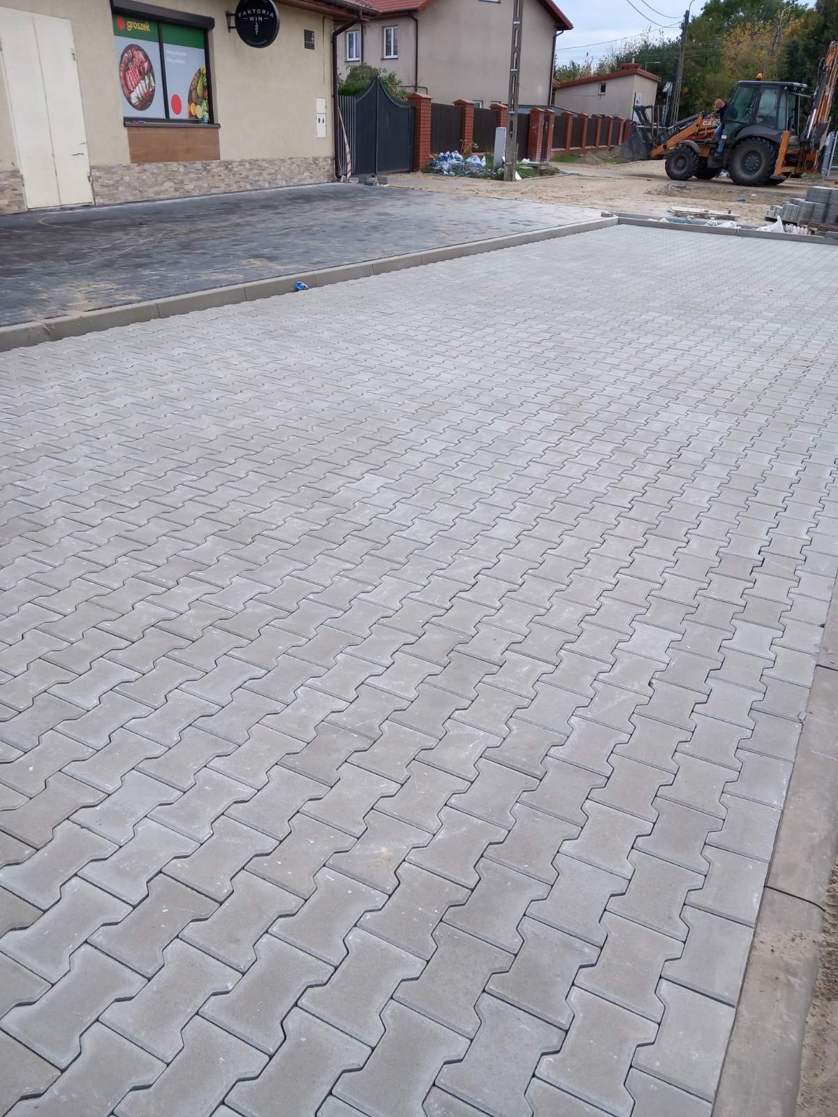 Ułożona szara kostka brukowa typu 'I' tworząca duży plac przed budynkiem z szyldem 'Kantoria WIN'. W tle widoczna koparko-ładowarka.