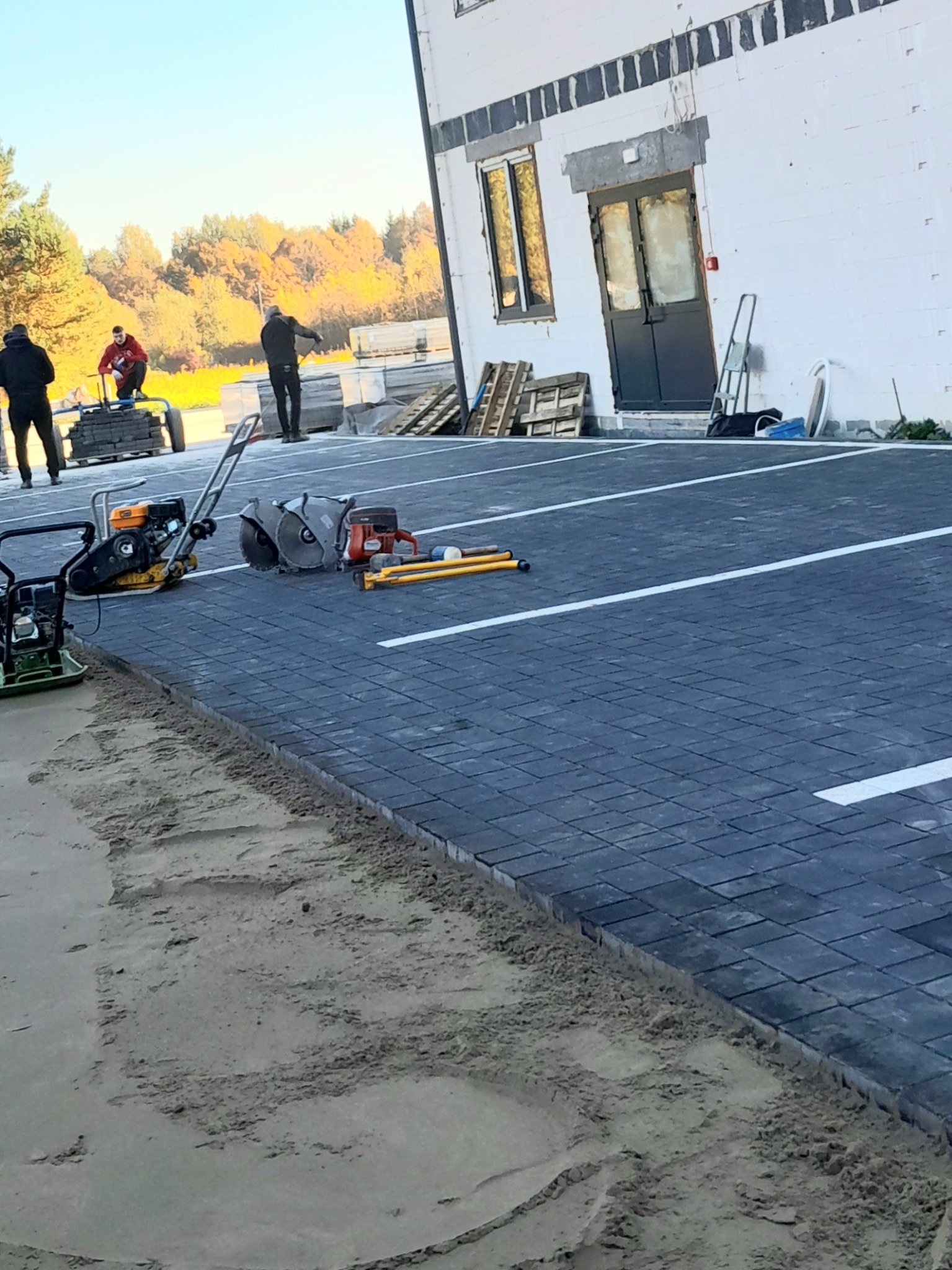 Niebieska kostka brukowa ułożona na parkingu z wyznaczonymi białymi liniami, widoczne narzędzia brukarskie i pracownicy w tle, budynek w trakcie wykończenia.