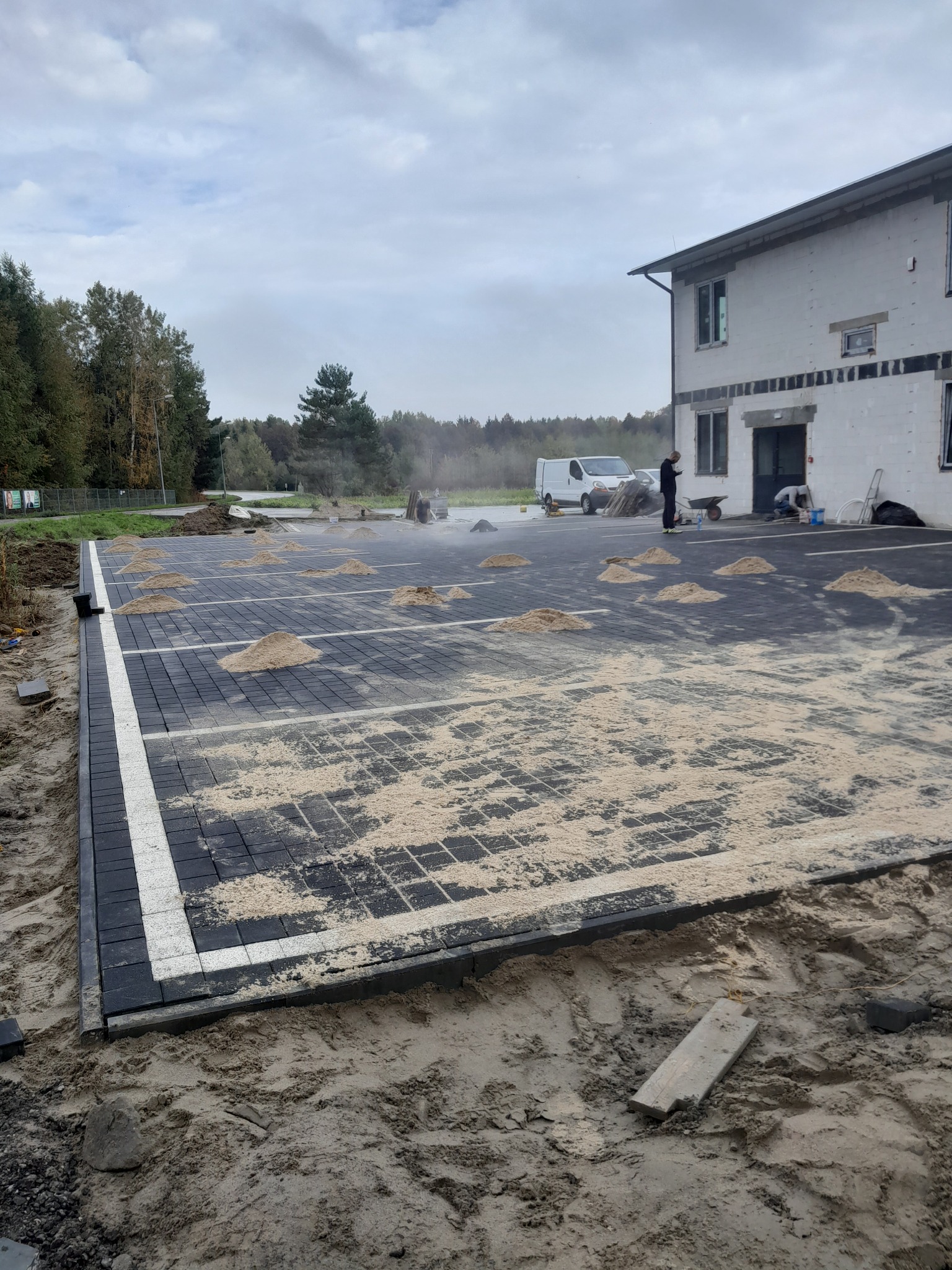 Częściowo ułożona, czarna kostka brukowa z białym pasem, przygotowana pod parking, z kopcami piasku na wierzchu, na tle surowego budynku i pracowników.