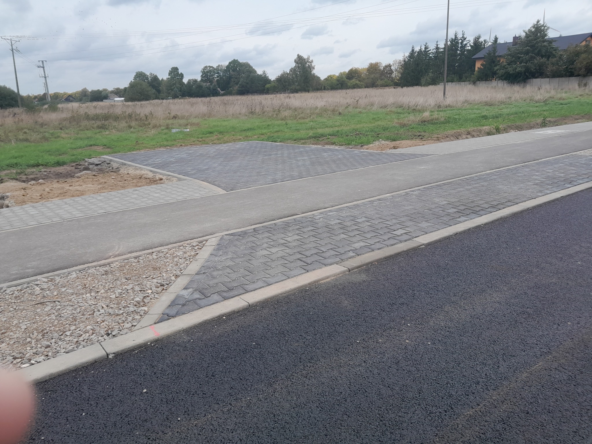 Nowa nawierzchnia z kostki brukowej i asfaltu na obrzeżach działki, widoczne przygotowane podłoże z piasku i kamieni, tło stanowią drzewa i pole.