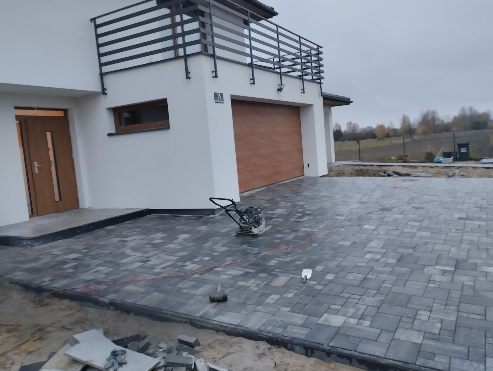 Nowo ułożona, szara kostka brukowa przed nowoczesnym domem z brązową bramą garażową. Widoczna ubijarka wibracyjna, młotek gumowy i łopatka do piasku. Częściowo ułożone obrzeża. W tle jesienny...