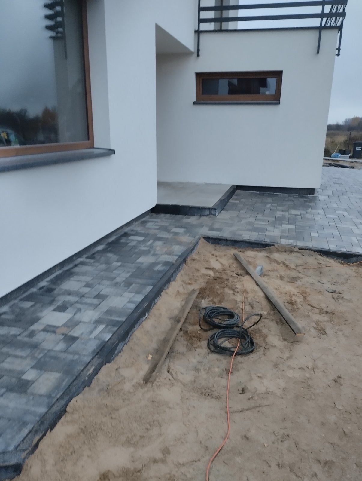 Fragment posesji w trakcie prac brukarskich: ułożona część ścieżki z szarej kostki, przygotowany teren z widocznymi kablami i drewnianymi listwami, biały budynek w tle.