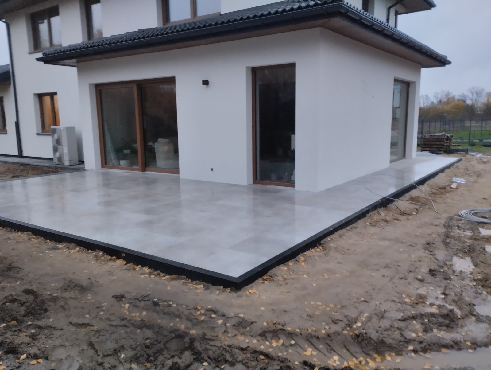 Nowoczesny taras z jasnych płyt ułożonych przy białym domu z dużymi oknami, obrzeże wykończone czarnym krawężnikiem, widoczne ślady prac ziemnych.
