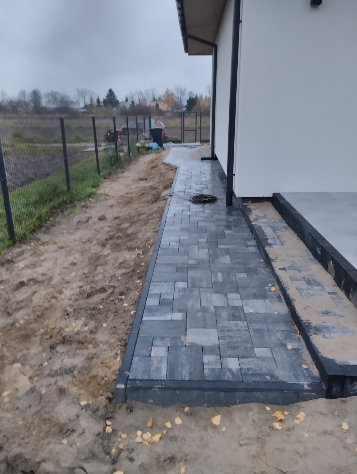 Ułożona, szara kostka brukowa tworząca ścieżkę wzdłuż domu w Warszawie, widoczna granica z ziemią i ogrodzeniem z siatki, pochmurny dzień.