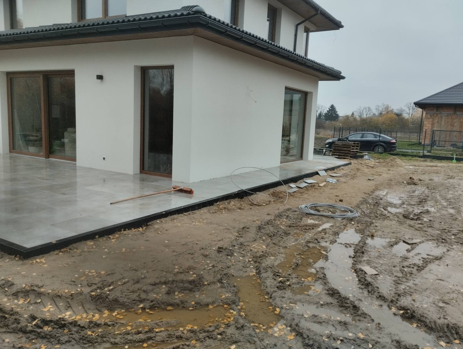 Nowoczesny taras z szarych płyt na zewnątrz domu, z widocznymi śladami błota i prac wykończeniowych na trawniku, w tle czarny samochód i ogrodzenie.