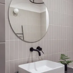 Nowoczesna łazienka w Warszawie z szarymi płytkami ułożonymi pionowo, okrągłe lustro z czarnymi elementami, biała umywalka i czarna bateria, uzupełniona małą rośliną w czarnej doniczce.