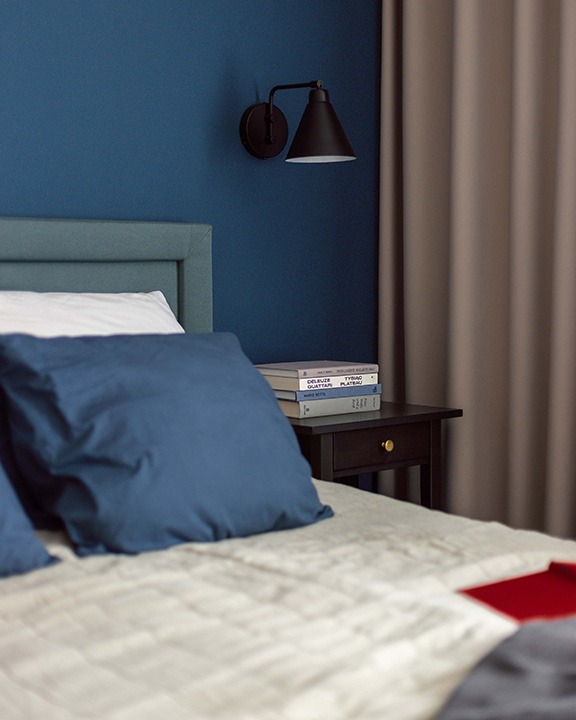 Sypialnia w odcieniach granatu i beżu: łóżko z niebieskimi poduszkami, czarna lampka nocna na ścianie, stolik z książkami i zasłony w tle.