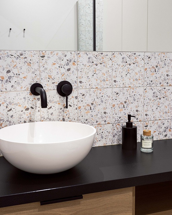 Nowoczesna łazienka z okrągłą, białą umywalką nablatową, czarną armaturą i blatem, oraz mozaikową ścianą z płytek w odcieniach szarości, bieli i pomarańczy.
