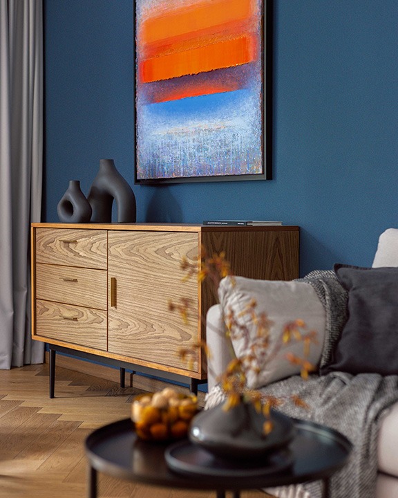 Nowoczesny salon z drewnianą komodą, abstrakcyjnym obrazem na niebieskiej ścianie i dekoracyjnymi wazonami