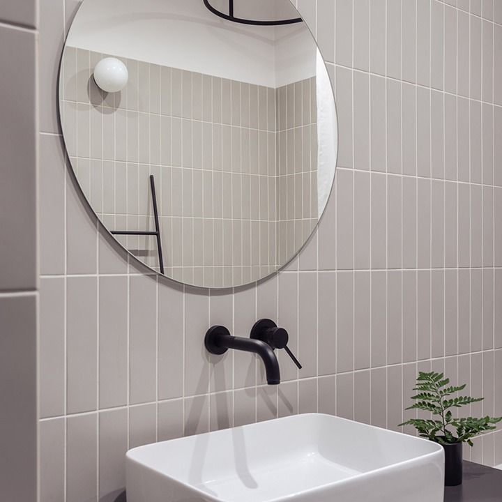 Nowoczesna łazienka w Warszawie z szarymi płytkami ułożonymi pionowo, okrągłe lustro z czarnymi elementami, biała umywalka i czarna bateria, uzupełniona małą rośliną w czarnej doniczce.