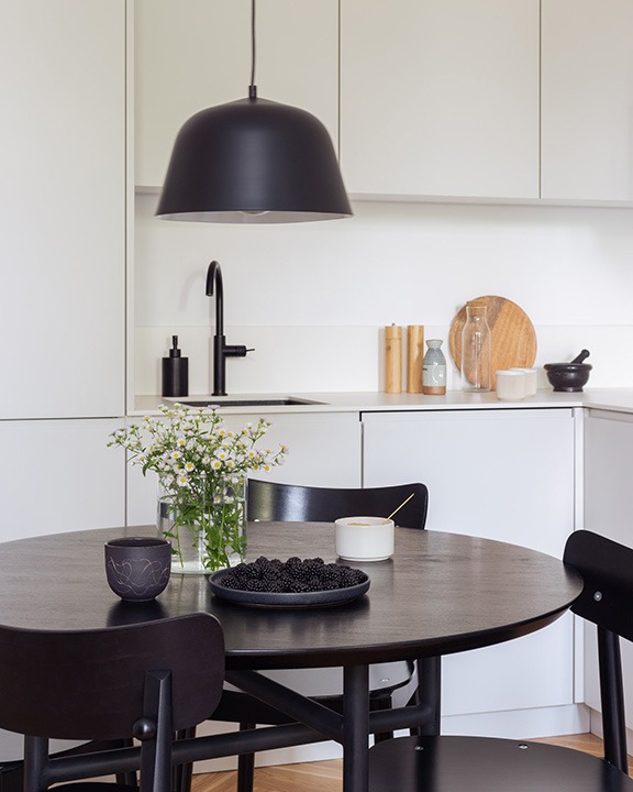 Nowoczesna kuchnia z białymi szafkami, czarną armaturą i okrągłym, ciemnym stołem. Na stole wazon z polnymi kwiatami, miseczka z jeżynami i czarna lampa wisząca w kształcie kopuły.