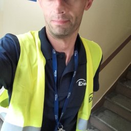 PNB Wrona euro-bud Olek Worona - Pracownik Enea w żółtej kamizelce odblaskowej i czapce z logo firmy, stojący na klatce schodowej z tabliczką 'Wejście do piwnicy'.