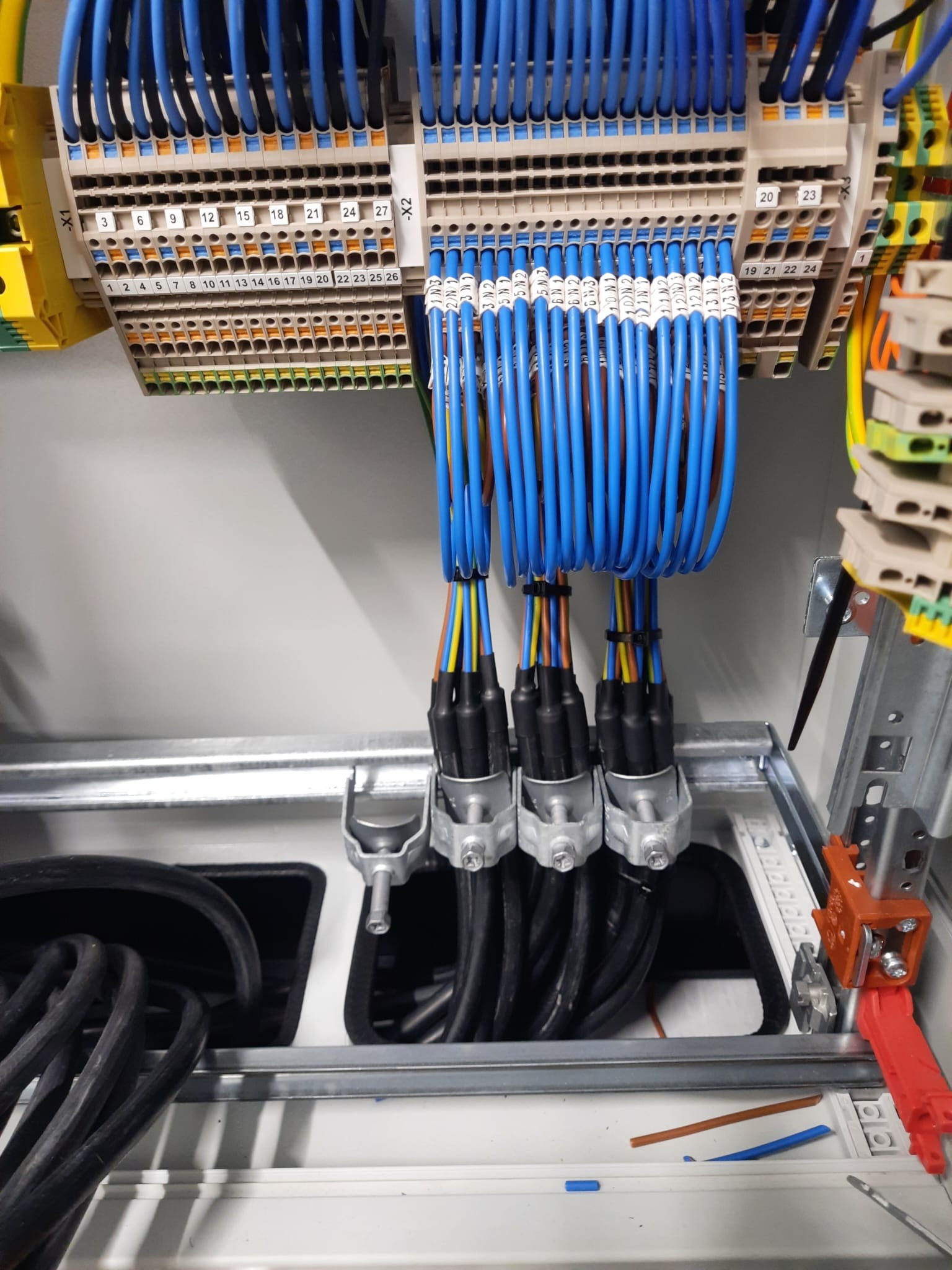 Szczegółowe ujęcie rozdzielnicy elektrycznej z podłączonymi, ponumerowanymi przewodami w kolorze niebieskim i czarnym, uporządkowanymi w metalowych uchwytach.