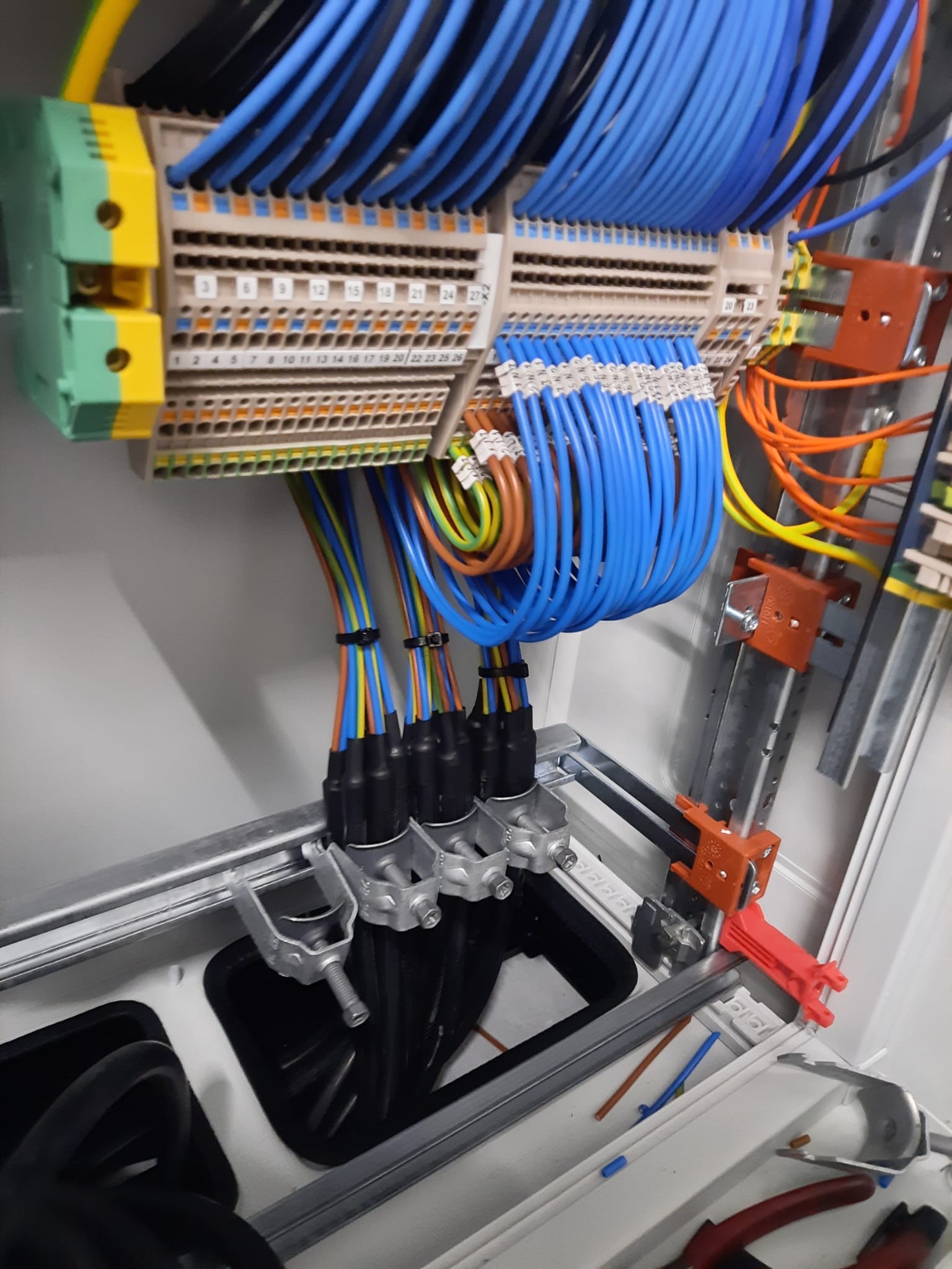 Panel elektryczny z uporządkowanymi kablami w kolorach niebieskim, żółtym, brązowym i zielonym, podłączonymi do listew zaciskowych. Widoczne metalowe szyny montażowe i narzędzia w dolnej części szafy.