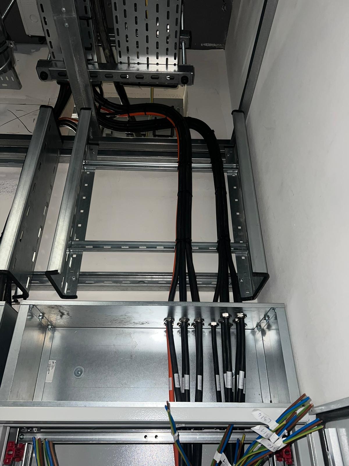 Metalowa korytka kablowa z poprowadzonymi przewodami elektrycznymi w czarnej izolacji, wchodzącymi do metalowej skrzynki rozdzielczej z widocznymi opisami kabli.