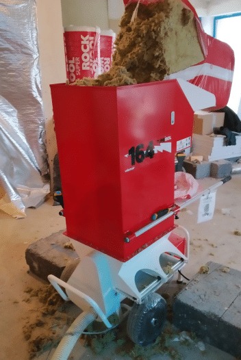 Czerwona maszyna do wdmuchiwania izolacji celulozowej z widocznym zasypanym włóknem, na tle folii paroizolacyjnej i materiałów budowlanych.