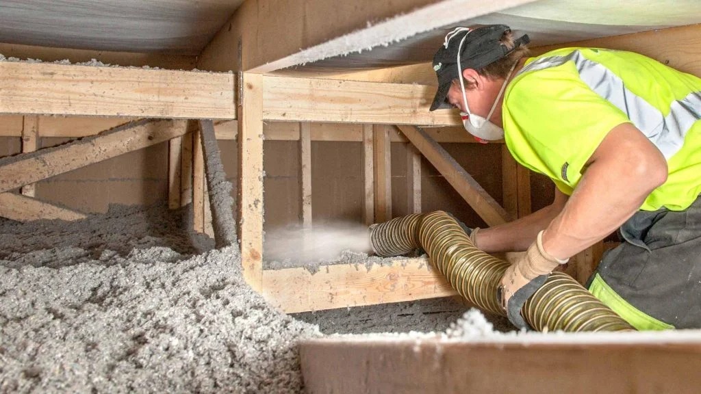 Pracownik w masce ochronnej aplikuje izolację z wełny mineralnej za pomocą węża do drewnianej konstrukcji poddasza, widoczne drewniane belki i warstwa izolacji na podłodze.