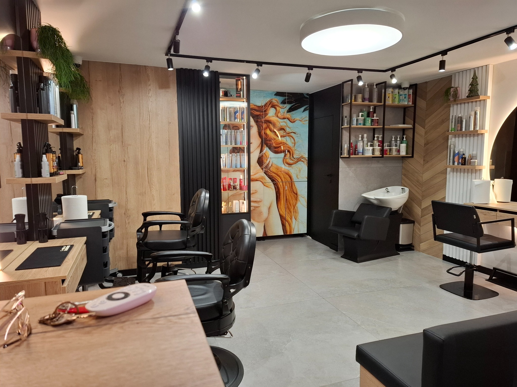 Nowoczesne wnętrze salonu fryzjerskiego z czarnymi fotelami, oświetleniem punktowym i dekoracyjnym panelem ściennym z wizerunkiem kobiety, widok z perspektywy recepcji z kluczami i okularami.