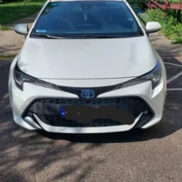 Biała Toyota Corolla Hybrid z polską tablicą rejestracyjną zaparkowana na asfaltowym parkingu w słoneczny dzień, widok z przodu, w tle zieleń drzew i budynek.