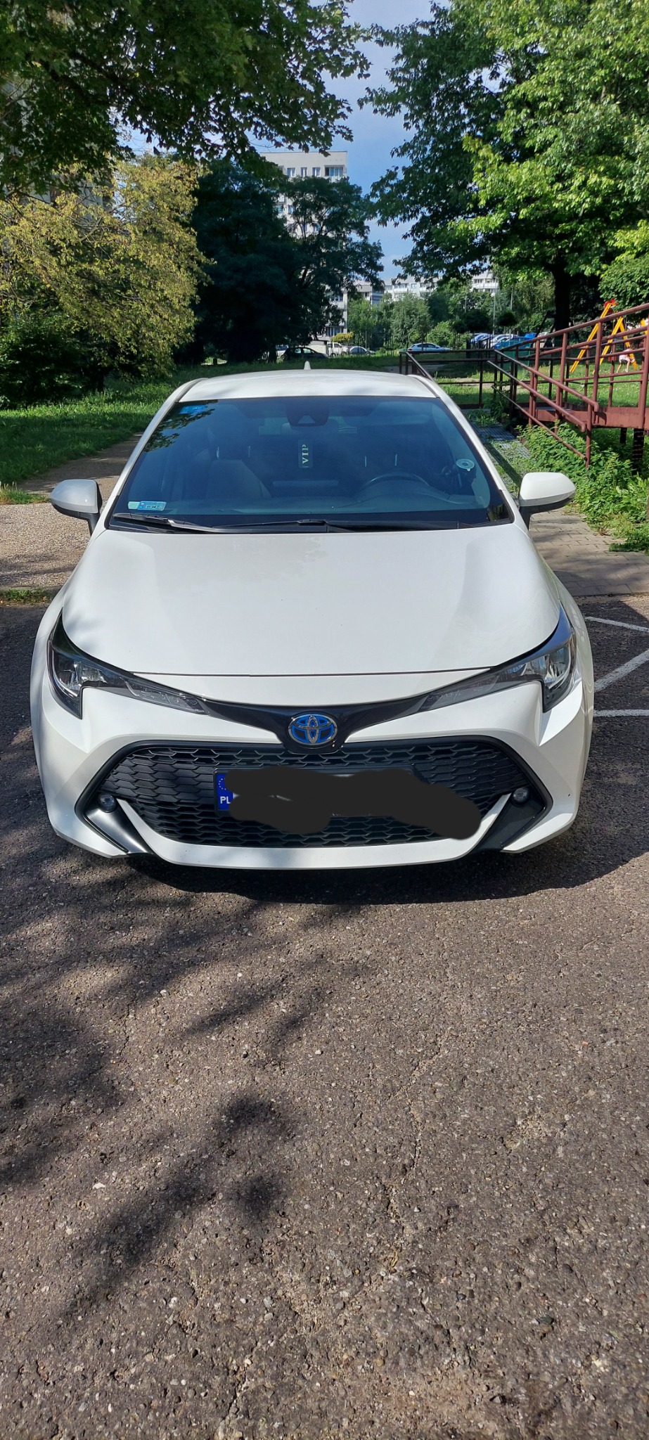 Biała Toyota Corolla Hybrid z polską tablicą rejestracyjną zaparkowana na asfaltowym parkingu w słoneczny dzień, widok z przodu, w tle zieleń drzew i budynek.