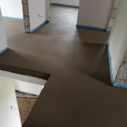 Świeżo wylana, gładka posadzka w surowym stanie, zabezpieczona niebieską taśmą przy ścianach, widoczne niedokończone fragmenty ścian i instalacje.