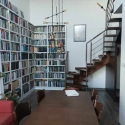 Przestronne wnętrze domu z biblioteką obejmującą całą ścianę, drewnianymi schodami z metalową barierką, nowoczesną lampą wiszącą i dużym drewnianym stołem z dokumentami.