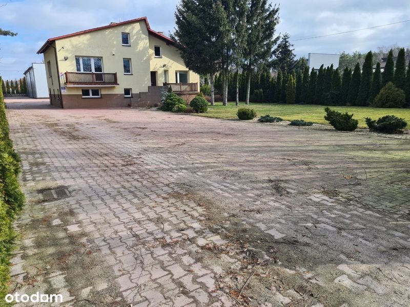 Żółty dom dwukondygnacyjny z balkonami, widok z podjazdu wyłożonego kostką, zielony trawnik i tuje w tle.