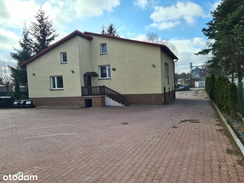 Dwukondygnacyjny budynek mieszkalny z brązowym dachem i schodami wejściowymi w Grudziądzu, otoczony brukowanym podjazdem i zielenią.