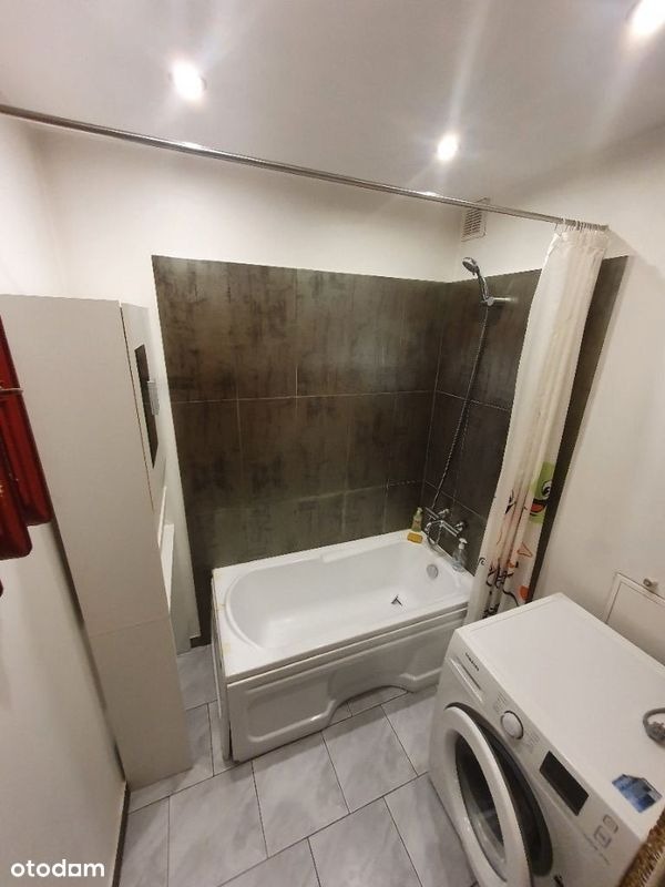 Wnętrze łazienki z wanną obudowaną płytkami, pralką i zasłonką prysznicową w rogu.