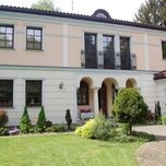 Elewacja dwukondygnacyjnego domu z beżową fasadą, czarnymi oknami i balkonem, otoczona zadbanym trawnikiem i zielenią.