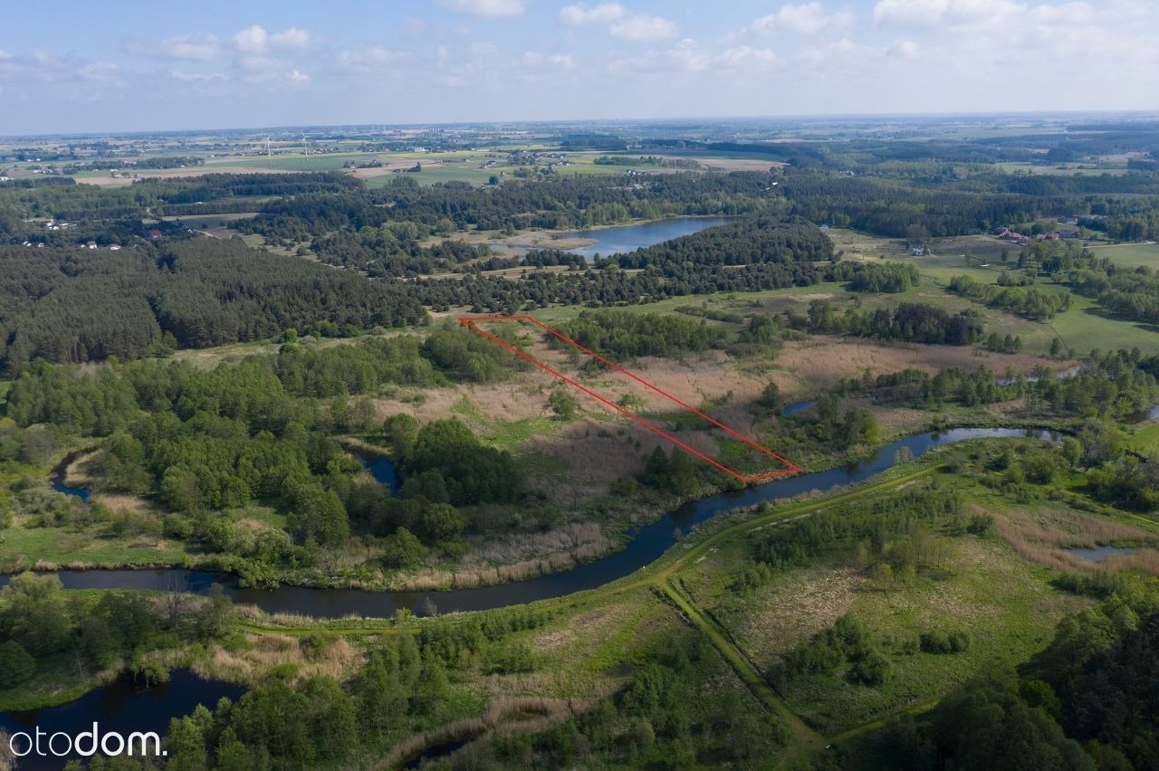 Zdjęcie z lotu ptaka przedstawia działkę przy rzece, zaznaczoną czerwoną linią, otoczoną zielenią i trzcinami.