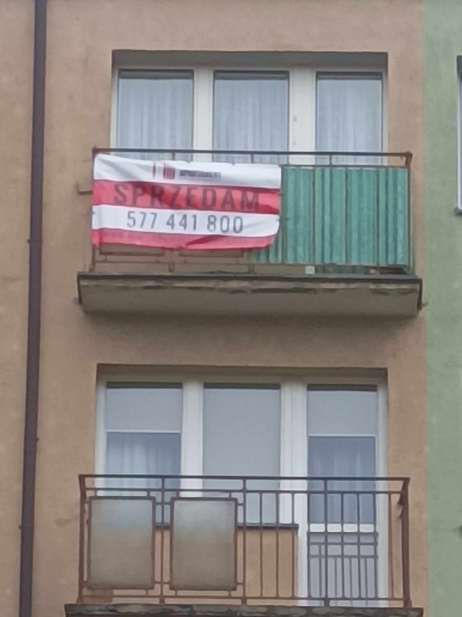 Balkon z wywieszoną reklamą 'SPRZEDAM' i numerem telefonu na tle bloku z oknami.
