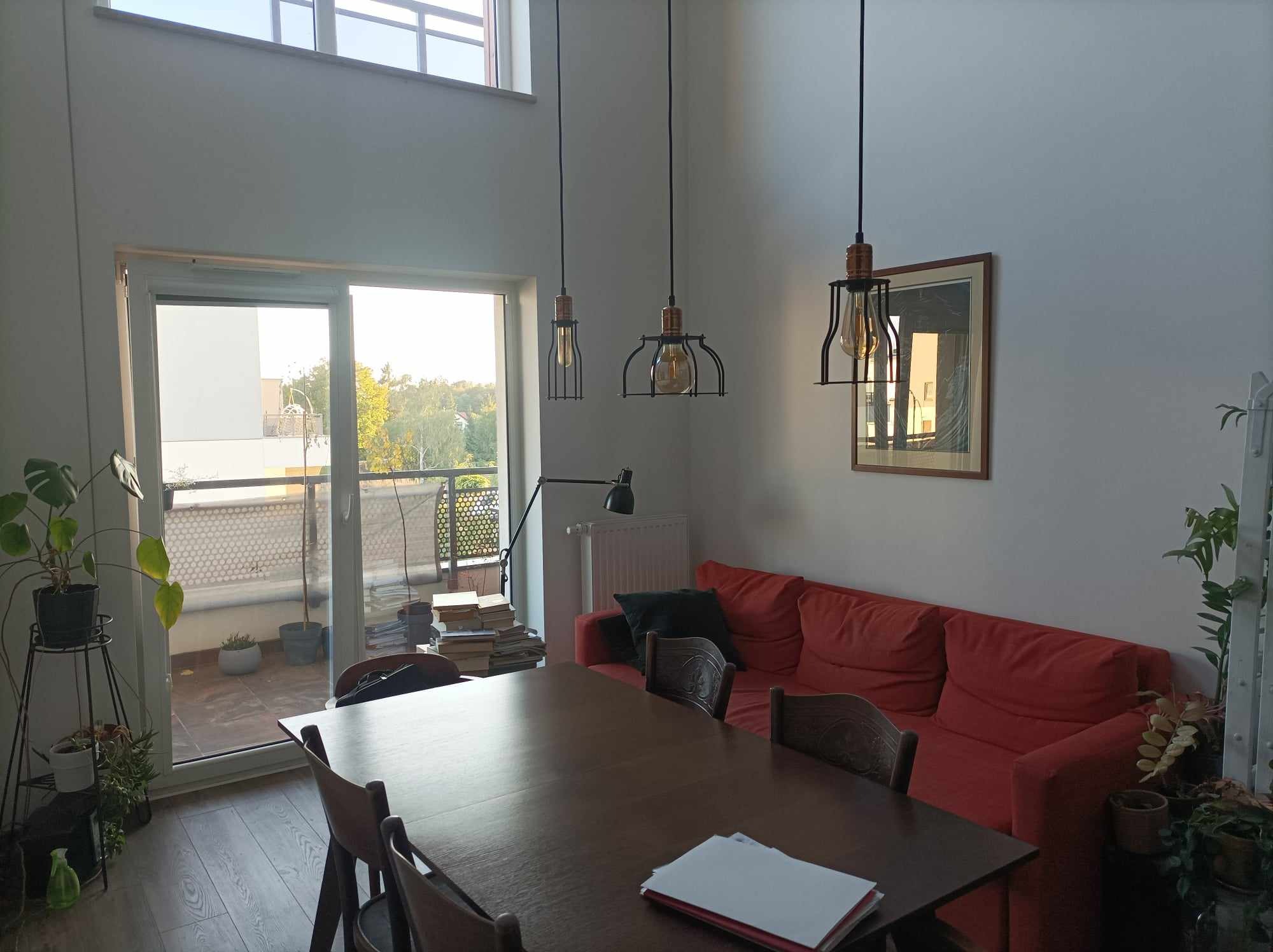 Jasne wnętrze mieszkania z wysokim sufitem, drewnianym stołem i czerwonym narożnikiem, oświetlone trzema wiszącymi lampami w stylu industrialnym, z widokiem na balkon i zieleń za oknem.