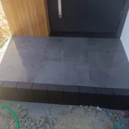 Wykończone kamienne schody wejściowe do domu z szarych płyt, obrzeża z czarnej kostki, widoczny fragment elewacji i zielony wąż ogrodowy.