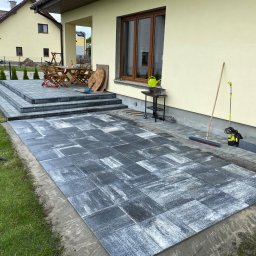 Antek Wojteczek - Niedokończony taras z szarych płyt betonowych o nieregularnej fakturze, z widocznymi narzędziami: szczotką i myjką ciśnieniową, oraz fragmentem trawnika z wężem ogrodowym.