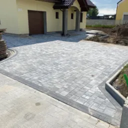 Nowoczesny podjazd z szarej kostki brukowej ułożonej we wzór jodełki, z obrzeżem z ciemniejszych kostek, przed domem we Wrocławiu. Widoczne palety i przygotowane podłoże pod dalsze prace.