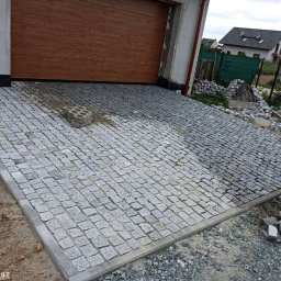 Antek Wojteczek - Nierównomiernie ułożona szara kostka granitowa przed garażem, widoczne pozostałości piasku i gruzu, w tle domy jednorodzinne.