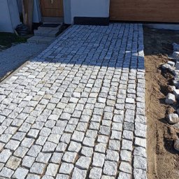 Antek Wojteczek - Świeżo ułożony chodnik z jasnoszarej kostki granitowej prowadzący do drzwi wejściowych domu, z widocznymi pozostałościami piasku na łączeniach i przygotowanym obszarem do dalszego układania.