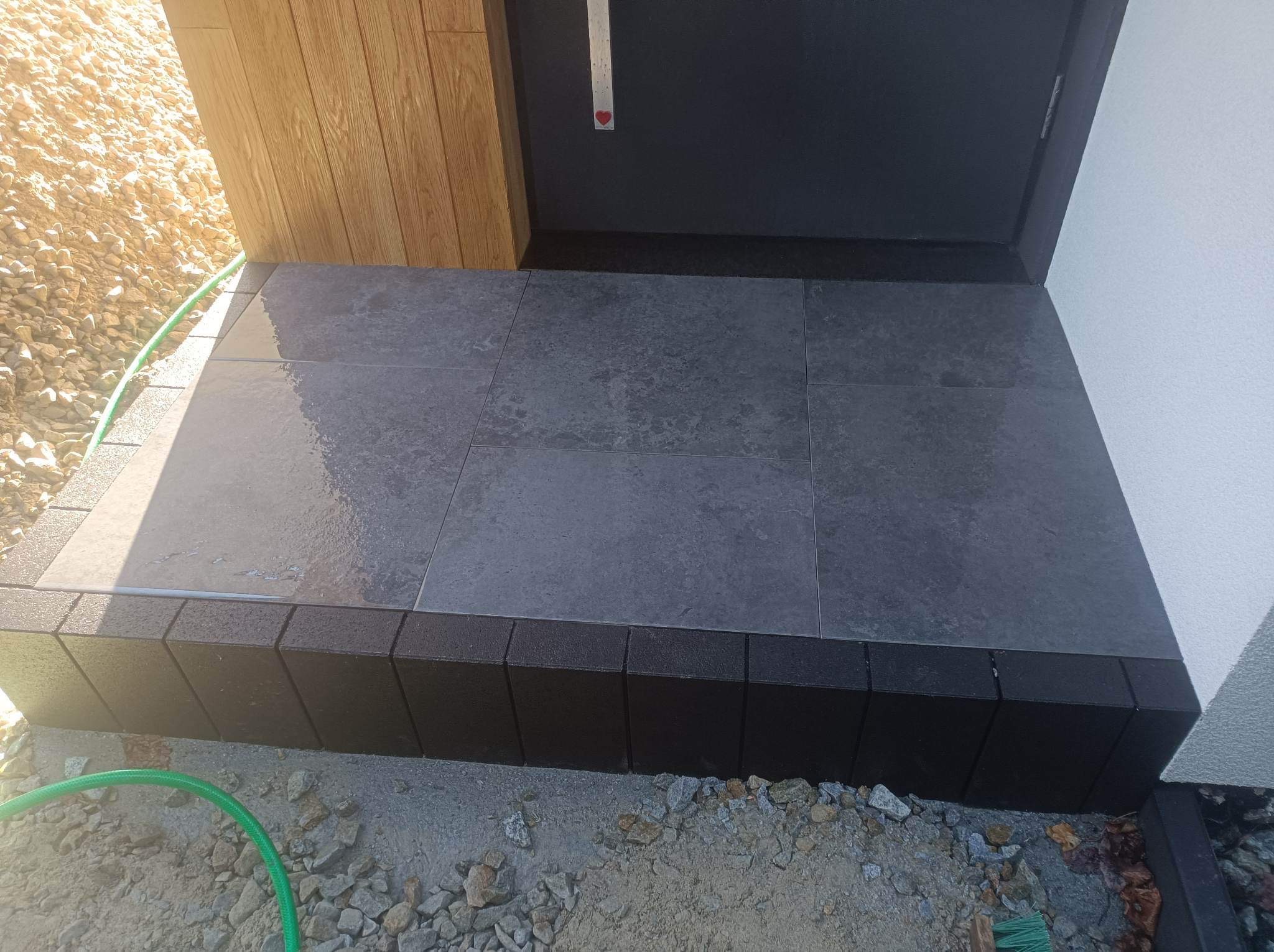 Wykończone kamienne schody wejściowe do domu z szarych płyt, obrzeża z czarnej kostki, widoczny fragment elewacji i zielony wąż ogrodowy.