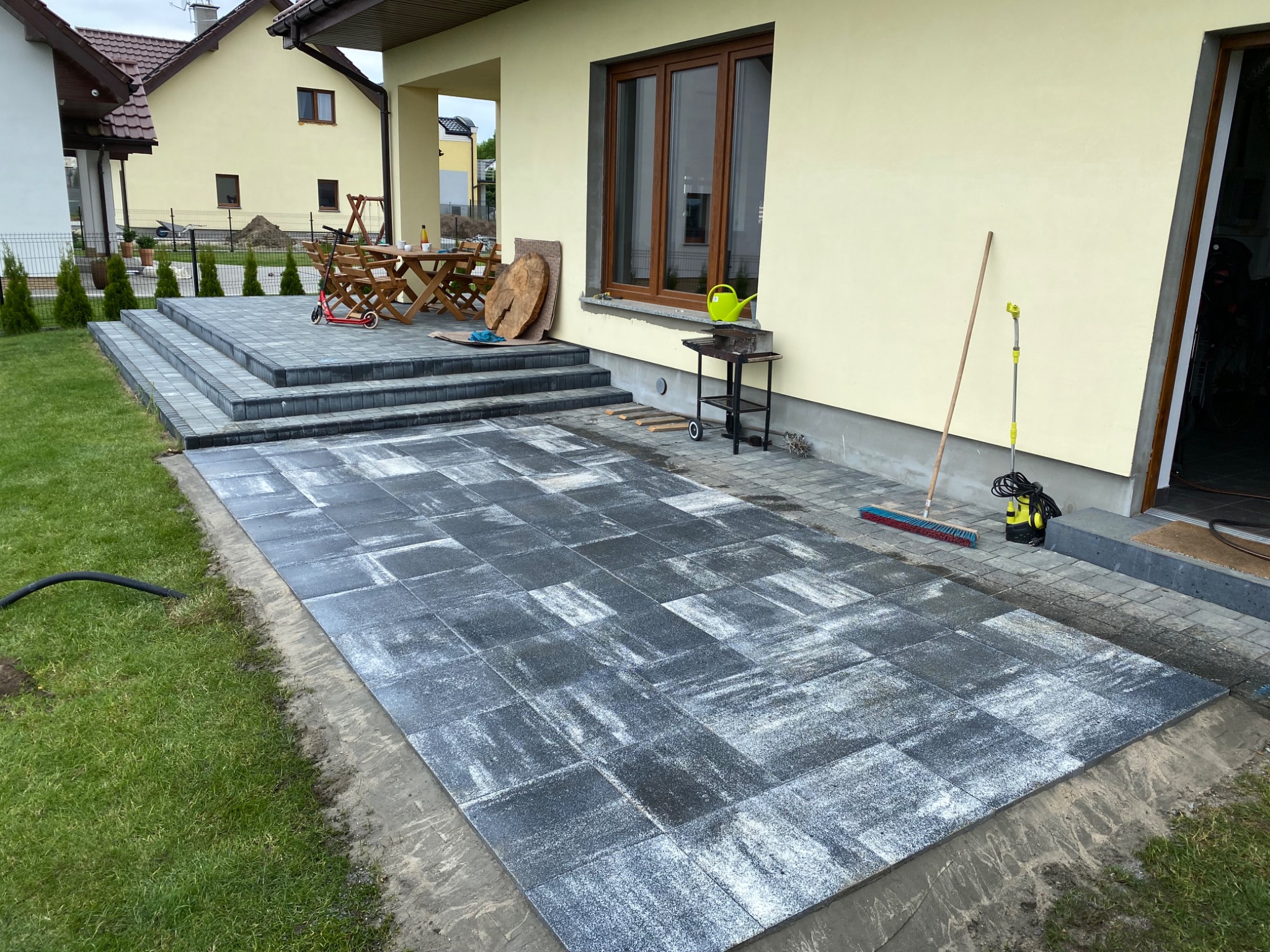 Niedokończony taras z szarych płyt betonowych o nieregularnej fakturze, z widocznymi narzędziami: szczotką i myjką ciśnieniową, oraz fragmentem trawnika z wężem ogrodowym.